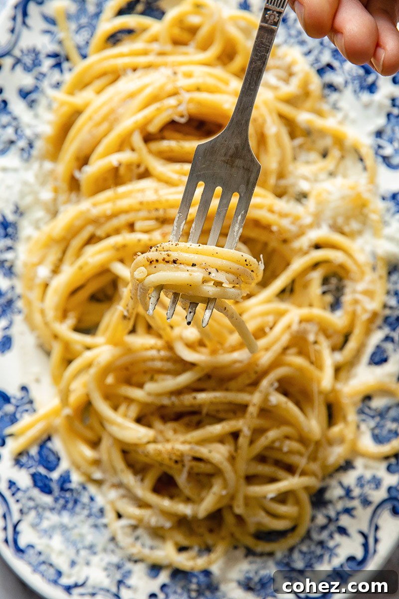 A few strands of bucatini Cacio e Pepe elegantly twirled on a fork, with a platter of pasta blurring in the background.