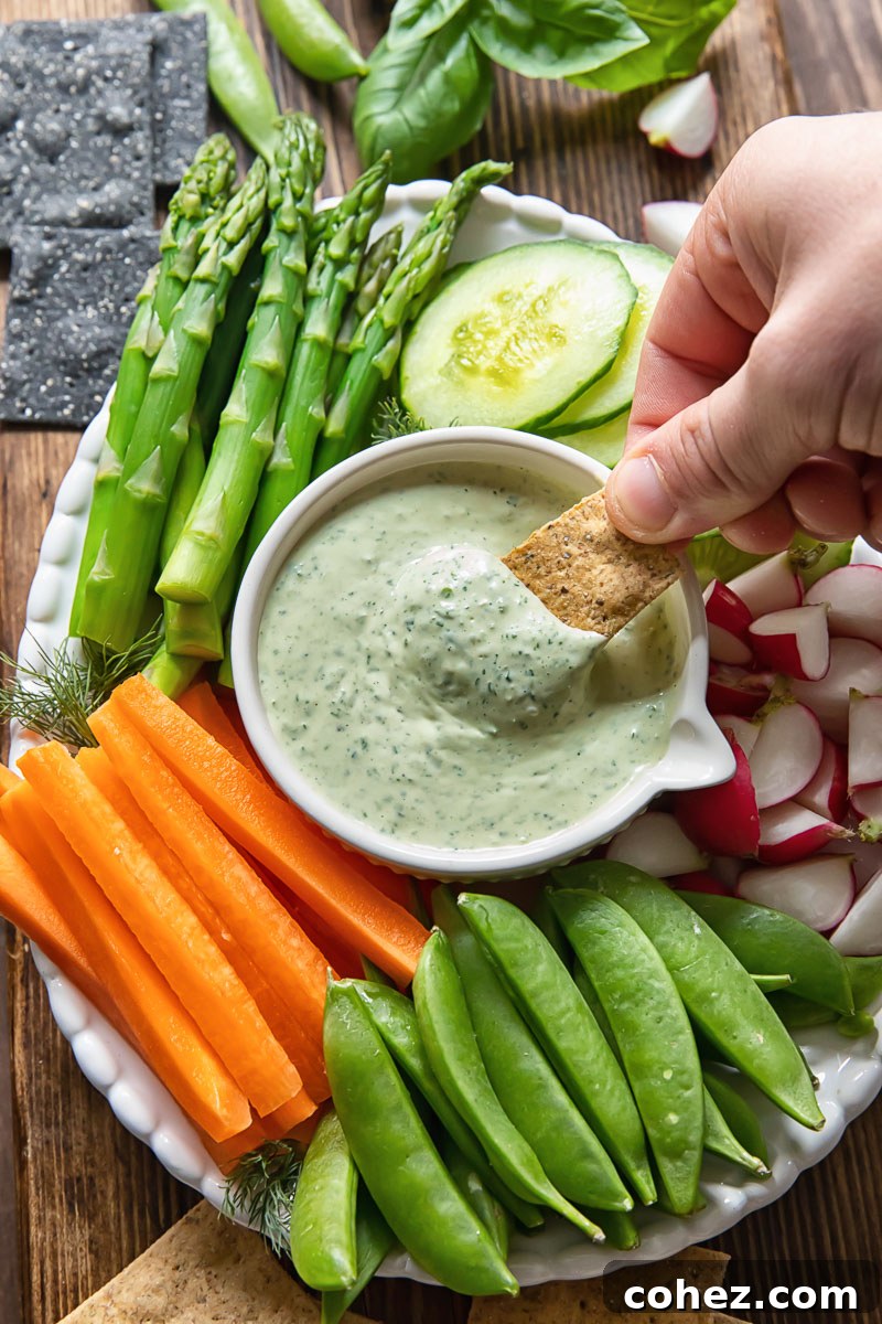 A crisp cracker being dipped into a bowl of green goddess dip, surrounded by a colourful array of fresh vegetables on a platter.