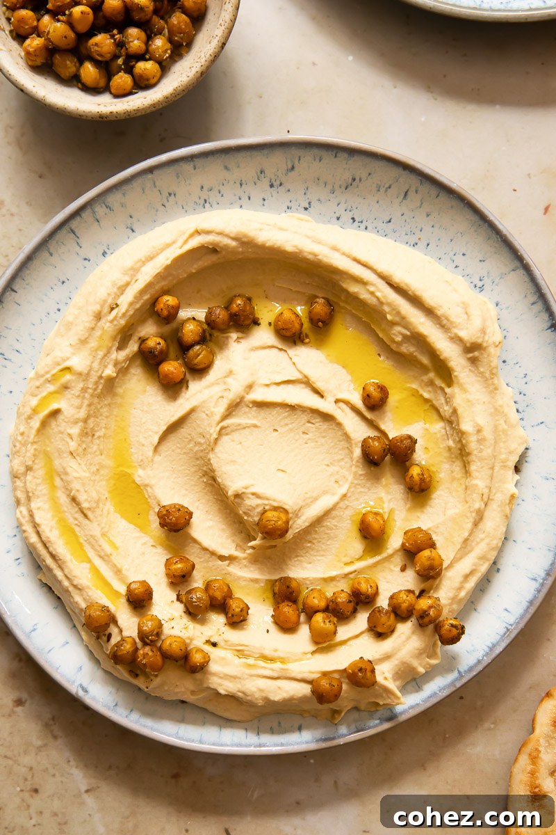 Close-up of freshly made hummus spread smoothly on a light blue ceramic plate, garnished with more chickpeas, a sprinkle of za'atar, and a generous drizzle of golden olive oil.