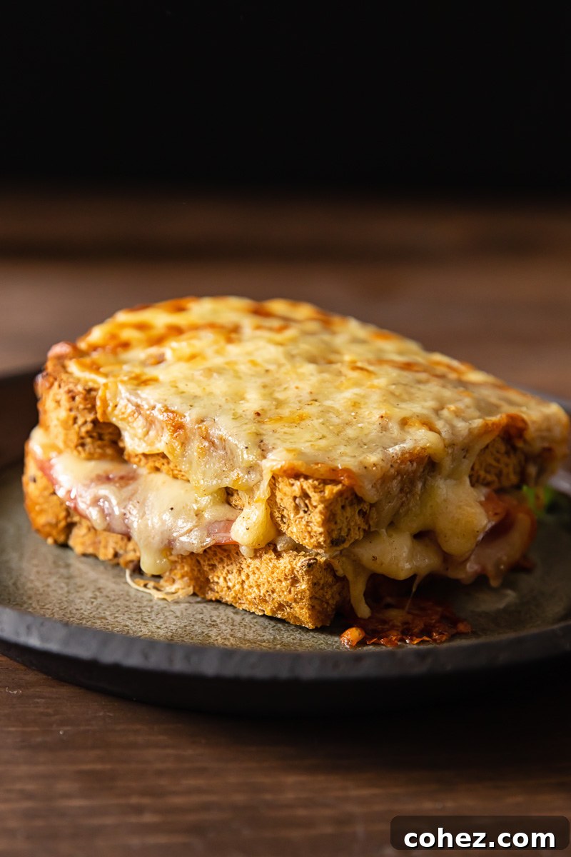 Ultimate Croque Madame 5 A side view of a golden-brown grilled Croque Madame sandwich, showcasing its perfectly toasted bread and melted cheese peeking from the edges, served on a rustic wooden board.