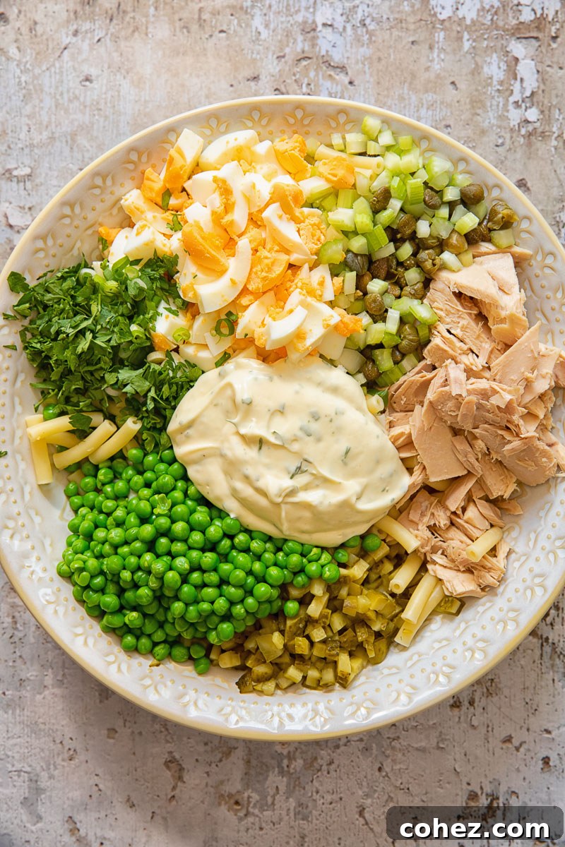 Process shot of tuna macaroni salad ingredients in a bowl before mixing, showing the individual components like pasta, tuna, peas, celery, and eggs.