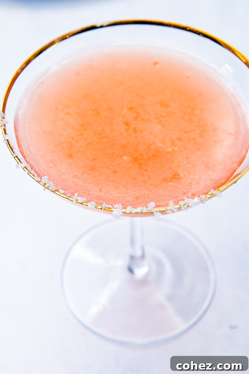 An overhead view of a beautifully prepared blood orange margarita in a salt-rimmed glass, garnished with a vibrant blood orange slice. The cocktail's deep reddish-orange hue is prominent against a neutral background, highlighting its appealing presentation.