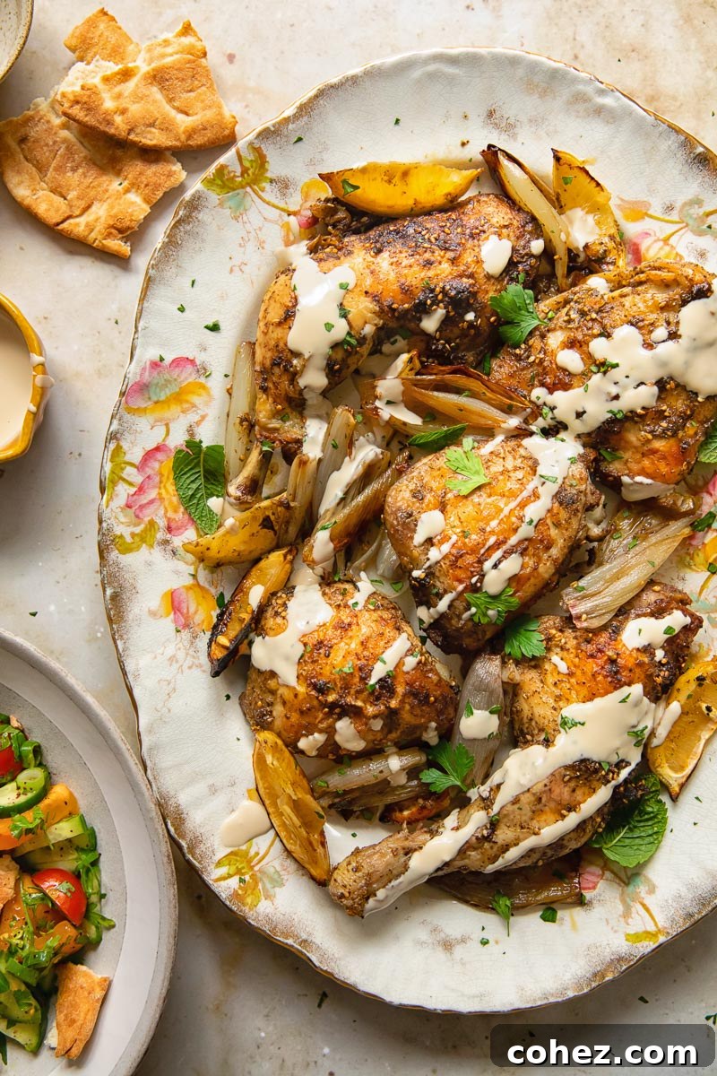 Top down view of tahini sauce drizzled over za'atar chicken with lemon and garlic on a platter, garnished with fresh herbs.