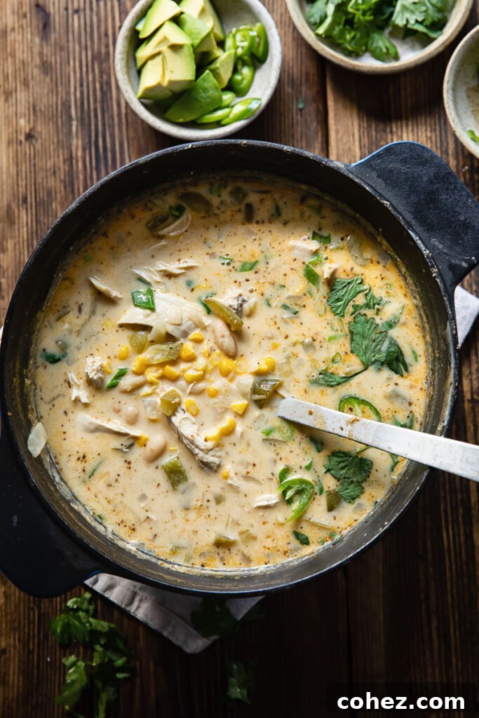 Top down shot of white chicken chili in a black pot next to a bowl of sliced avocado and cilantro