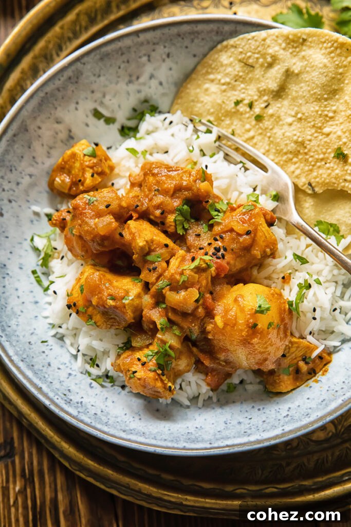 A close-up of a serving of chicken curry with potatoes, accompanied by rice and a papadam, invitingly presented in a bowl.