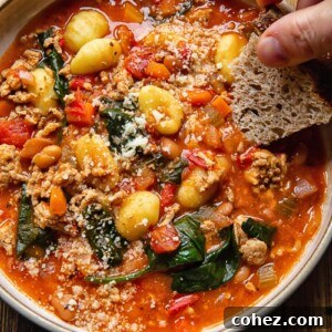 A steaming bowl of Firecracker Sausage Gnocchi Soup with a hand dipping a piece of crusty bread into it.
