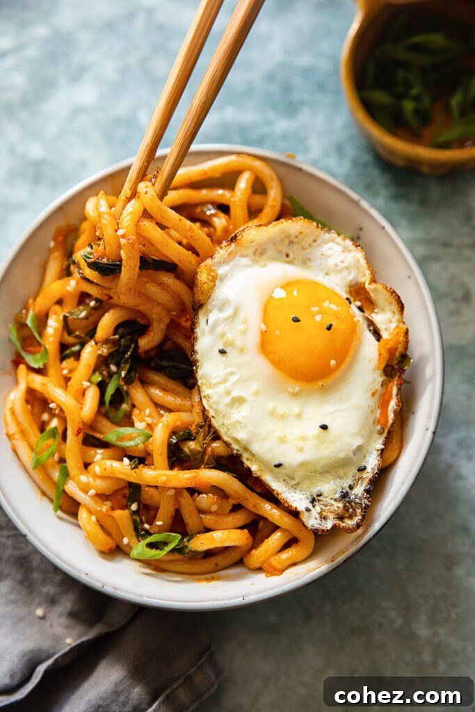 A beautifully presented bowl of kimchi noodles topped with a fried egg and garnished with fresh green onions and sesame seeds, ready to eat with chopsticks.