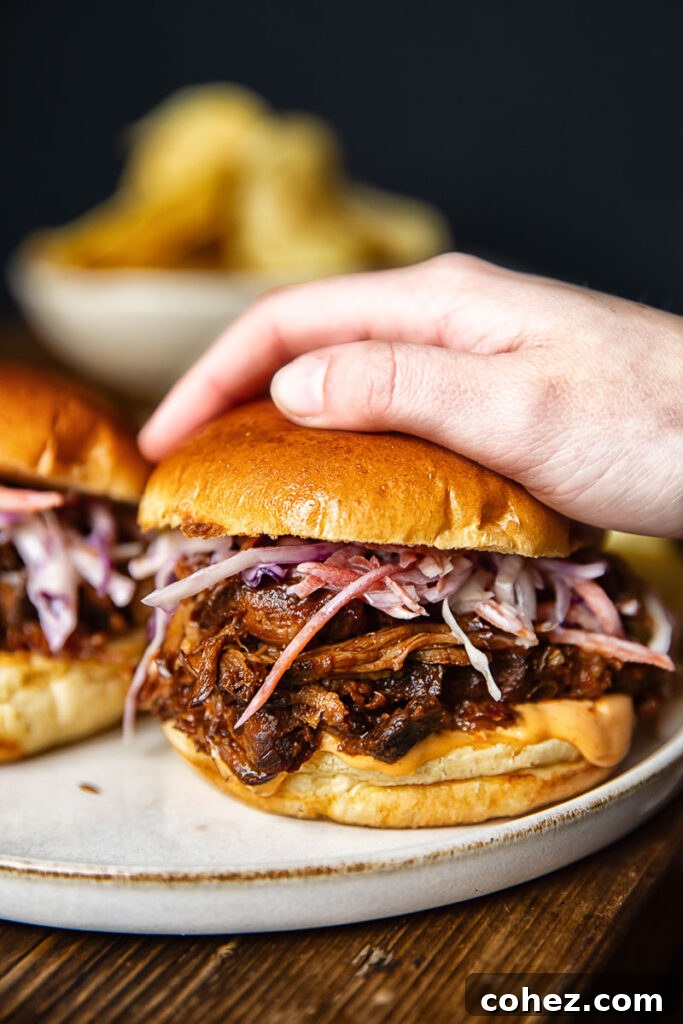 Side shot of someone picking up a pulled pork sandwich off of a plate next to another sandwich