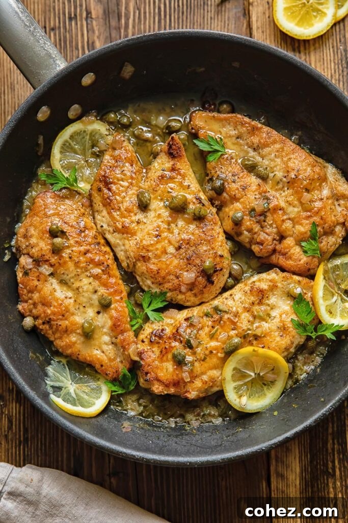 Top down view of chicken breasts in a pan, coated in piccata sauce with lemon slices and capers