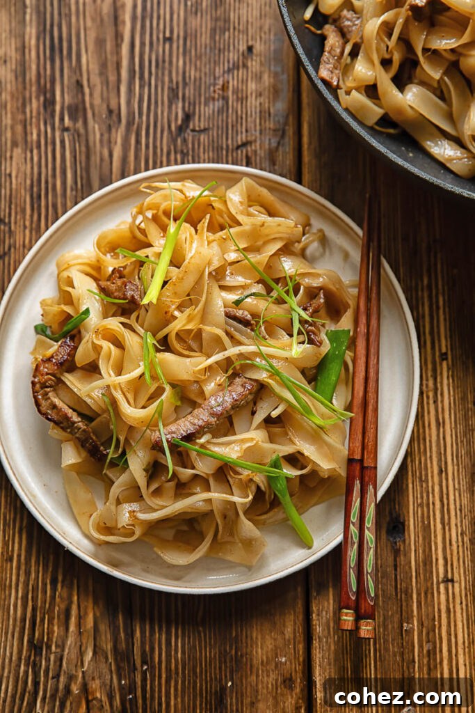 Top down shot of chow fun noodles topped with beef and green onions on a plate with chop sticks