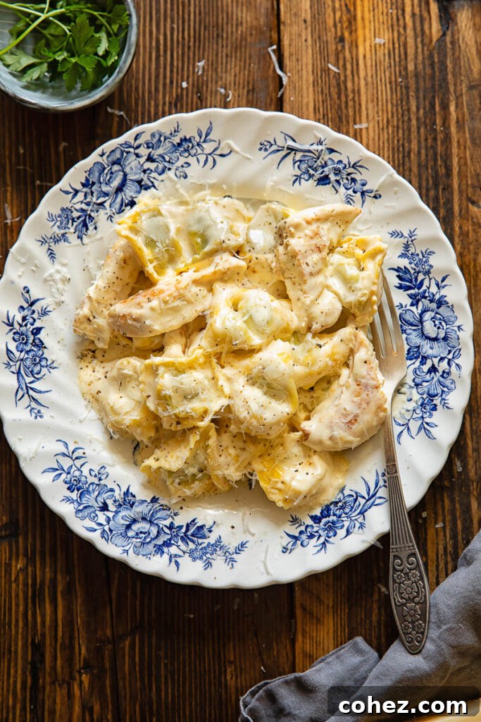 Top down shot of a fork in a bowl of chicken tortellini alfredo next to a bowl of green herbs