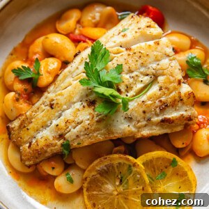Close up shot of fried cod with lemon butter sauce in a bowl on top of crushed cherry tomatoes and butter beans