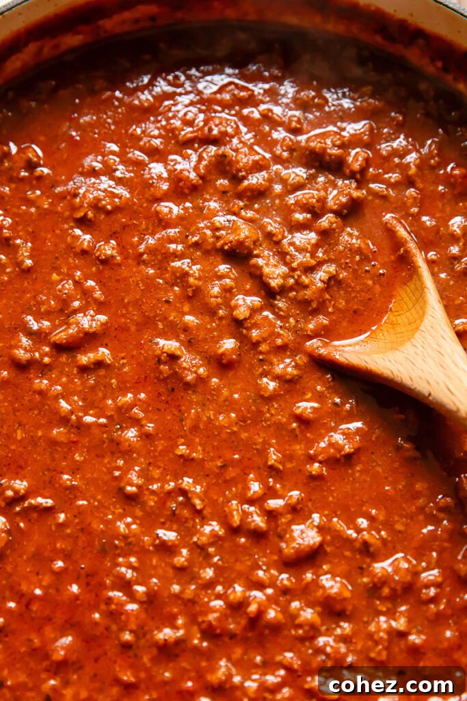 Close up of a vibrant beef and tomato sauce, enriched with 'nduja, simmering in a deep pan with a wooden spoon, showcasing its rich texture and color.