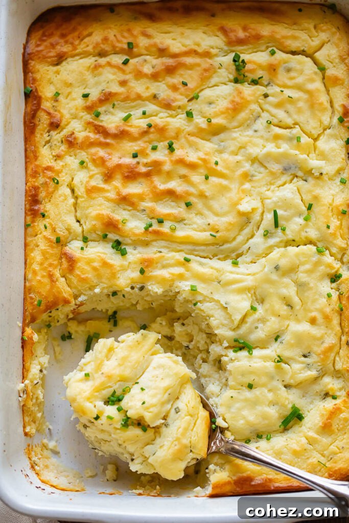 Golden Potato Cheese Cloud 3 A top-down view of a rectangular casserole dish filled with golden-baked potato soufflé, with a serving spoon taking a generous portion from the dish.