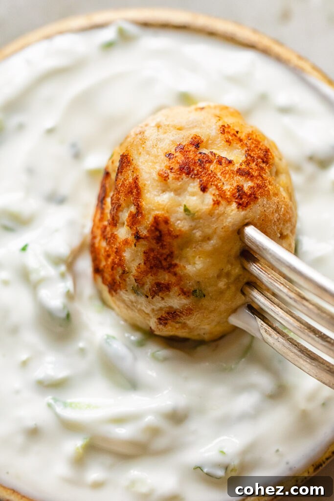 Close-up shot of a single chicken feta meatball being dipped into a bowl of homemade tzatziki, showing its perfectly browned exterior.
