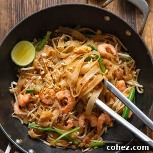 Wok with Pad Thai, with tongs removing a portion, next to a lime and peanuts
