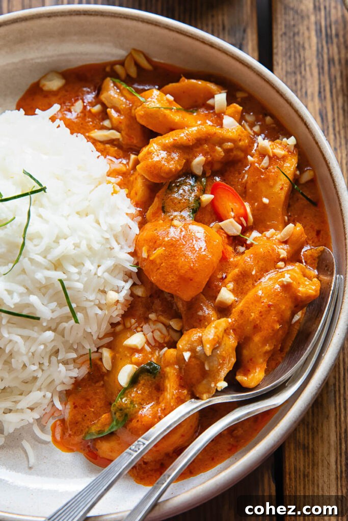 Close-up view of a bowl of Panang Chicken Curry next to a ball of jasmine rice, with a spoon and fork ready to serve.