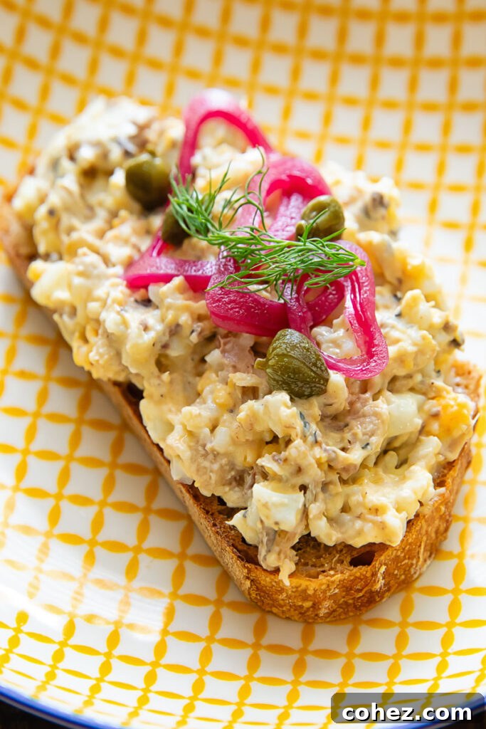 Toast on a yellow striped plate topped by a sardine mixture, capers, pickled onion and dill.